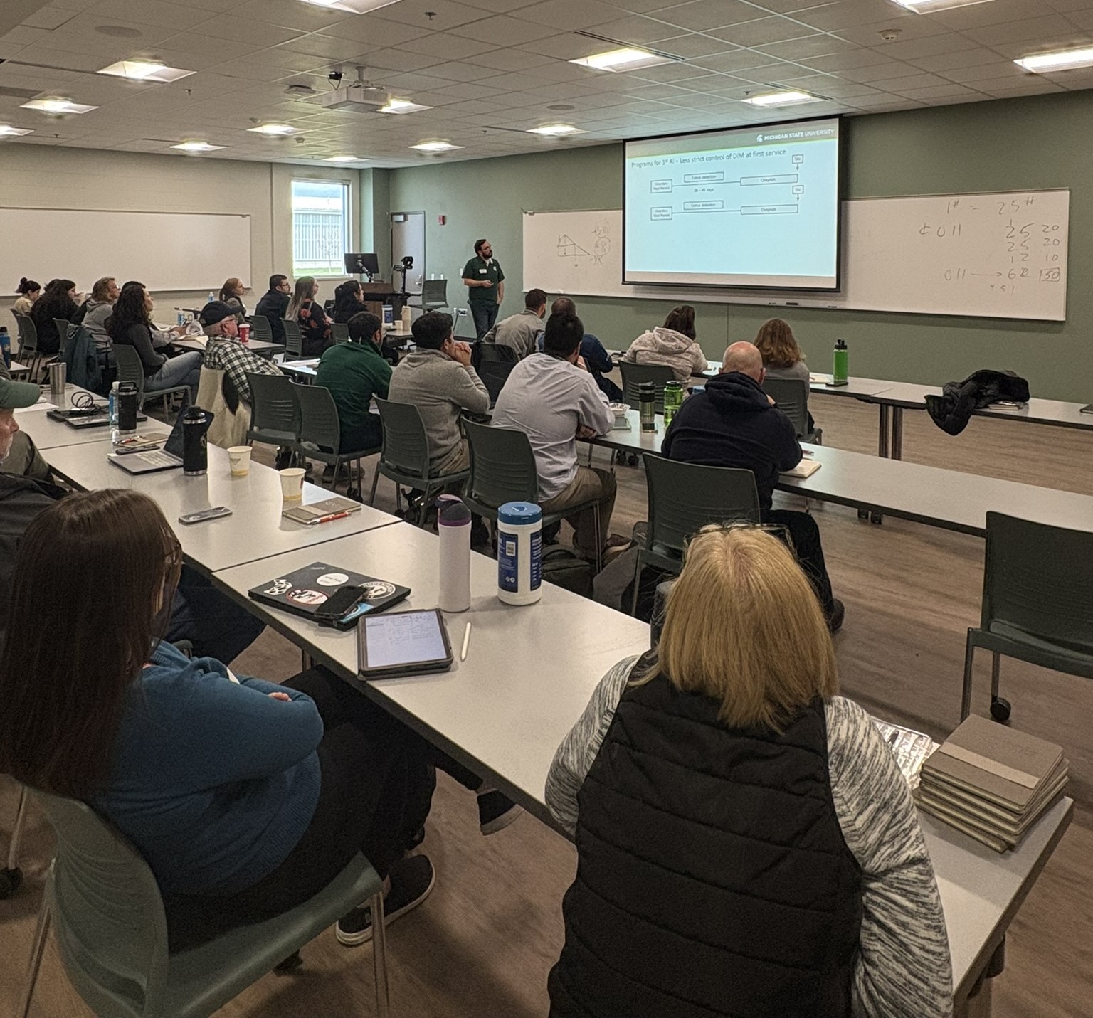 People sit at desks in a classroom, watching a presentation given at the head of the room.